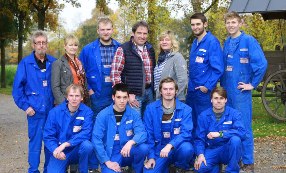 Groepsfoto van medewerkers in blauwe overalls, buiten in de natuur.